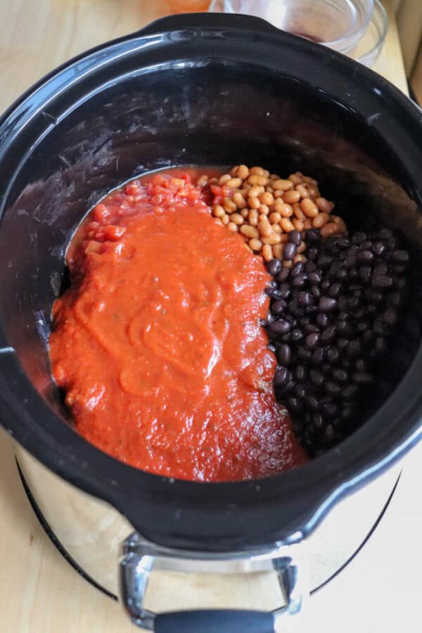 An easy crockpot chili awaits in a slow cooker brimming with diced tomatoes, baked beans, black beans, and tomato sauce. Each ingredient is uncooked and neatly arranged in sections, ready to transform into a comforting meal.
