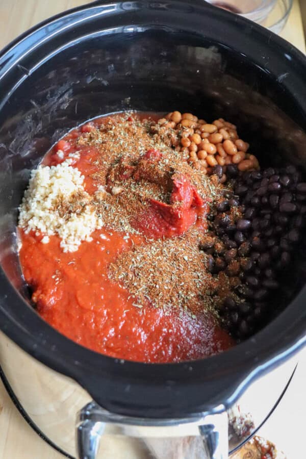 A slow cooker filled with ingredients for easy crockpot chili, including black beans, baked beans, crushed tomatoes, minced garlic, tomato paste, and various spices sprinkled on top.