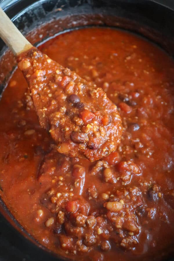 A wooden spoon holds up a portion of thick, hearty crockpot chili inside a slow cooker. This easy chili is rich and red, filled with ground meat, beans, and chopped tomatoes, showcasing its chunky texture and savory aroma.