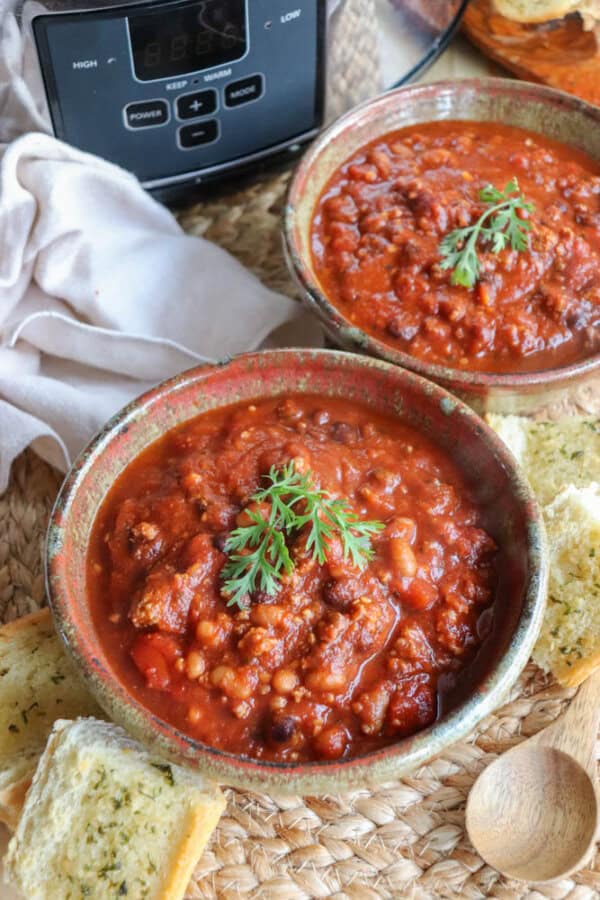 Two bowls of easy crockpot chili, topped with fresh herbs, rest on a woven mat. Slices of bread are nearby. A slow cooker and wooden spoon are visible in the background. The hearty chili is packed with beans and meat, offering a comforting meal.
