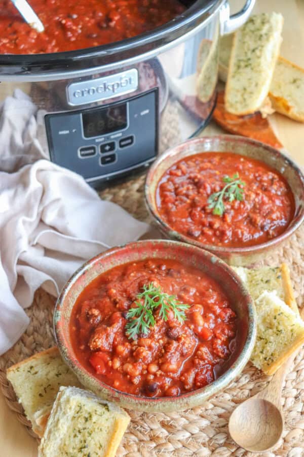 Two bowls of hearty, easy Crock-Pot chili topped with parsley sit on a woven mat, surrounded by slices of garlic bread. A Crock-Pot filled with more chili is in the background, with a wooden spoon and a napkin nearby.