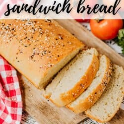A loaf of sandwich bread with sesame and poppy seeds, sliced on a wooden board. Red and white checkered cloth nearby, with tomatoes and greens in the background. Text overlay reads 5-Ingredient Sandwich Bread.