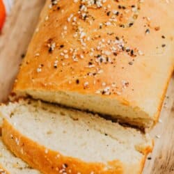 A loaf of homemade sandwich bread sprinkled with seeds rests on a wooden board. Several slices are cut, showing a soft, inner texture. Red and white kitchen cloth in the background. Text overlays the image.