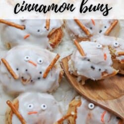 Cinnamon buns topped with white icing to resemble melted snowmen, featuring candy eyes, orange candy noses, and pretzel stick arms.