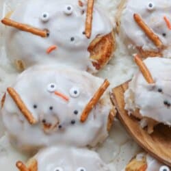 Cinnamon rolls decorated to look like snowmen with white icing, candy eyes, orange candy noses, and pretzel sticks for arms on a baking tray.
