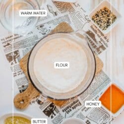 A flat lay image of ingredients for baking bread. Includes labeled warm water, yeast, flour in a bowl, honey, butter, and salt, arranged on a wooden board over newspaper.