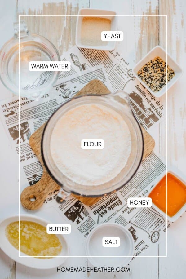 A flat lay image of ingredients for baking bread. Includes labeled warm water, yeast, flour in a bowl, honey, butter, and salt, arranged on a wooden board over newspaper.