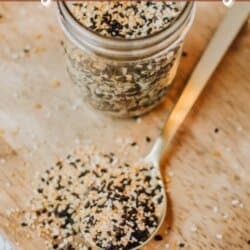 A wooden board holds a jar filled with homemade bagel seasoning. A gold spoon beside it is scooping some seasoning. The background has printed text, with HOMEMADE bagel seasoning written at the top and homemadeheather.com at the bottom.