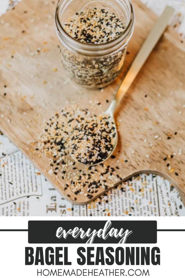 A jar and spoon filled with bagel seasoning sit on a wooden board. Some seasoning is scattered around the spoon. The text reads Everyday Bagel Seasoning and homemadeheather.com at the bottom.