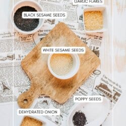 Overhead view of various seeds and seasonings in bowls on a wooden board, labeled as black sesame seeds, white sesame seeds, poppy seeds, garlic flakes, and dehydrated onion, placed on newspaper sheets.