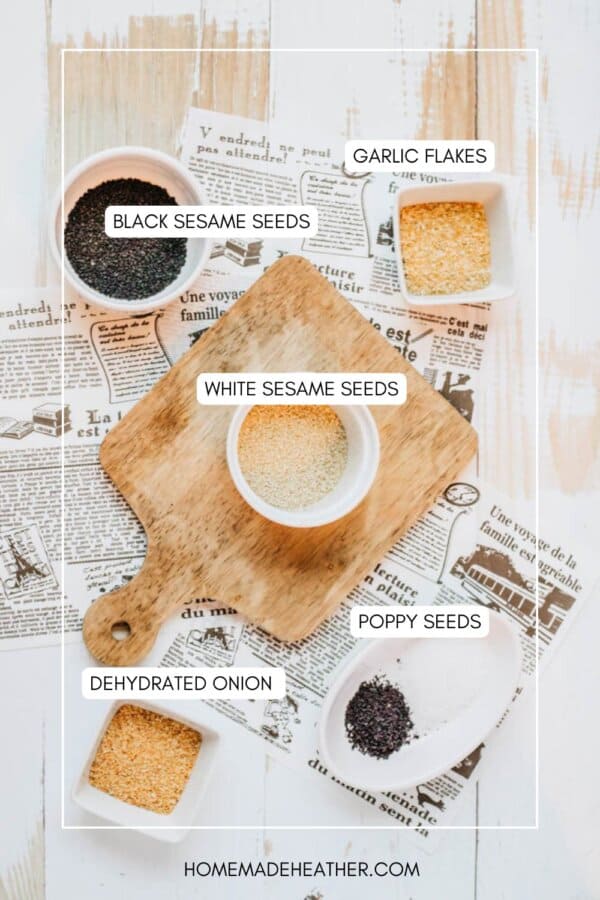 Overhead view of various seeds and seasonings in bowls on a wooden board, labeled as black sesame seeds, white sesame seeds, poppy seeds, garlic flakes, and dehydrated onion, placed on newspaper sheets.