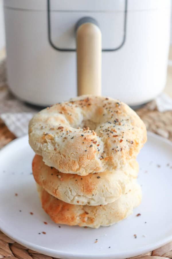 A stack of three homemade gluten-free bagels with a golden crust and sprinkled with seeds, placed on a white plate. A modern air fryer sits in the background, adding to the kitchens charm.