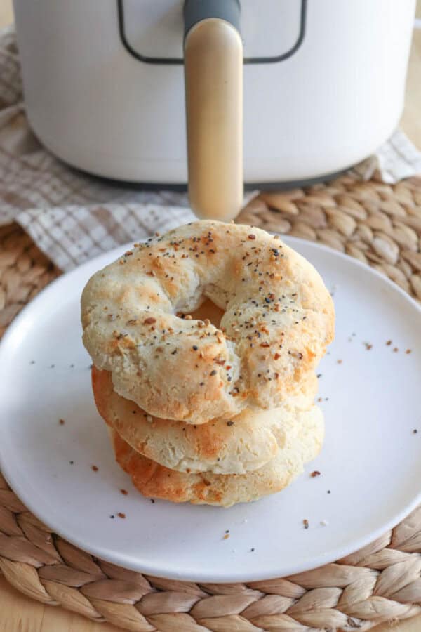 A stack of three homemade gluten-free bagels, sprinkled with seasoning, sits on a white plate. The plate rests on a woven placemat. In the background is an air fryer with a gold handle.
