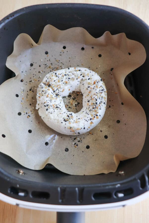 A gluten-free bagel, topped with savory seasoning, rests on parchment paper inside an air fryer basket.