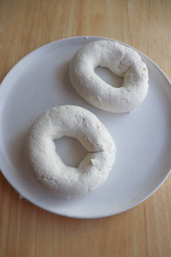 Two raw, gluten-free bagels crafted from dough sit on a round, white plate. The plate rests on a wooden surface. Perfect for an air fryer, these bagels are ready to transform into golden perfection.