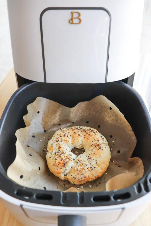 A gluten-free everything bagel is being air fried on parchment paper inside an air fryer basket. The bagel is topped with seeds and spices, showcasing a crispy golden texture.