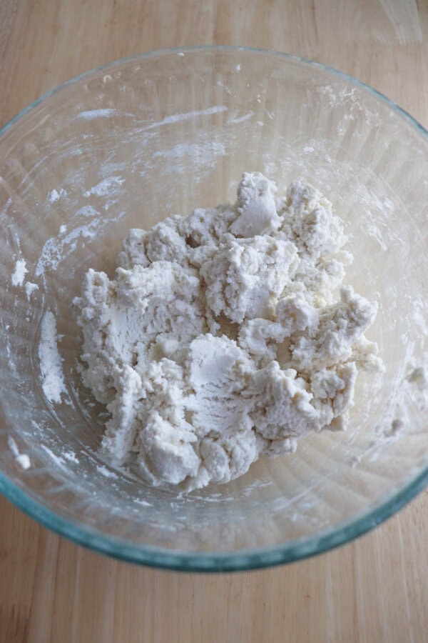 A glass bowl with a mixture of crumbled white dough sits on a wooden surface, the beginnings of gluten-free bagels being kneaded to perfection. Soon, theyll transform into golden delights in the air fryer.