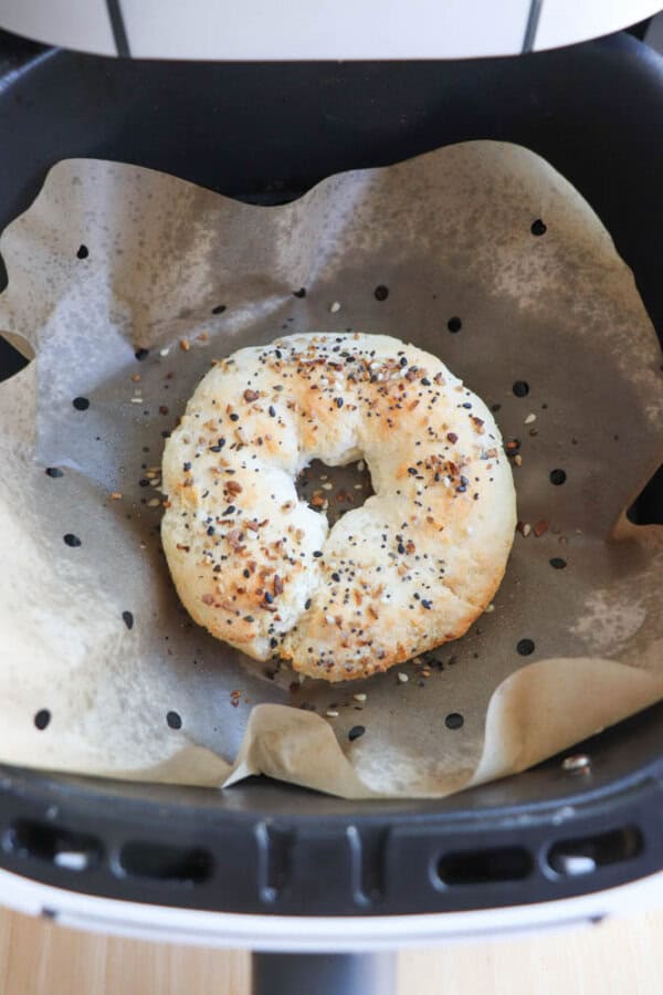 A gluten-free, freshly baked bagel topped with sesame and poppy seeds sits on parchment paper inside an air fryer basket.