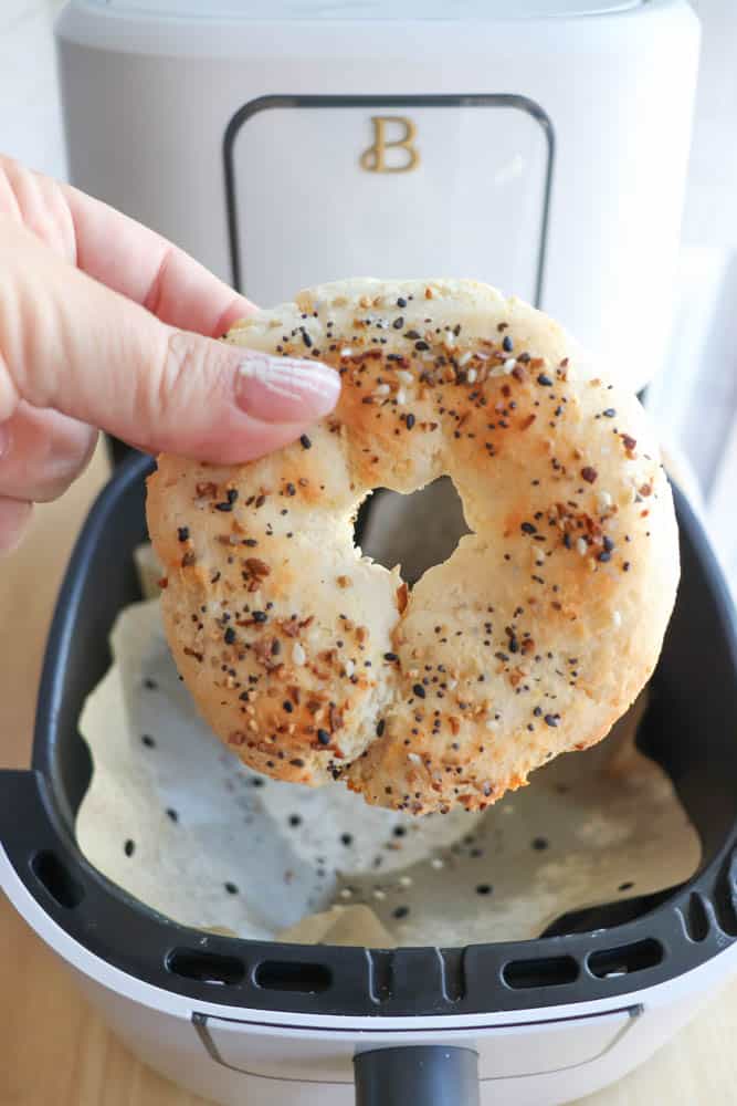 A hand holds a freshly baked gluten-free everything bagel over an air fryer basket. The bagel boasts a mix of seeds and spices. In the background, theres a sleek white air fryer with the letter B on it, perfect for achieving that crispy finish.