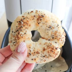 Close-up of a hand holding a freshly made gluten-free everything bagel in front of an air fryer. The bagel is golden brown with seasoning, and the persons fingernails are painted with white stripes.