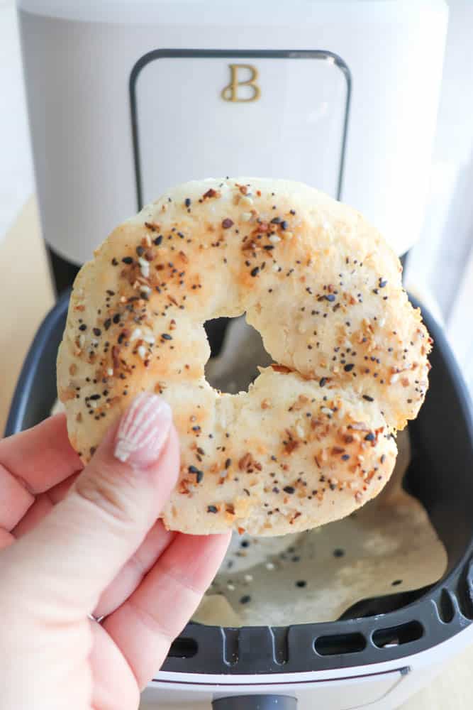 Close-up of a hand holding a freshly made gluten-free everything bagel in front of an air fryer. The bagel is golden brown with seasoning, and the persons fingernails are painted with white stripes.