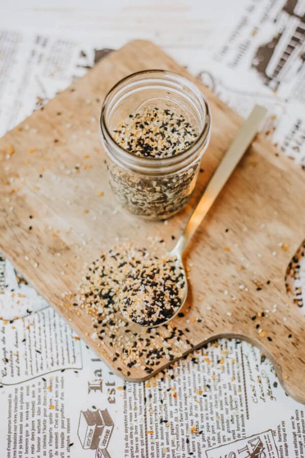 On a wooden cutting board, a glass jar filled with homemade everything bagel seasoning, featuring black and white seeds, stands invitingly. A spoon rests nearby, cradling some of the flavorful mix, while more seeds are scattered across the board atop newspaper clippings.