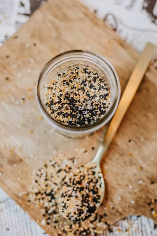 A jar filled with a black, white, and brown grainy mixture of homemade everything bagel seasoning sits on a wooden board. A golden spoon beside it holds a small amount of this savory blend. Some of the seasoning is spilled on the board, adding a touch of rustic charm.