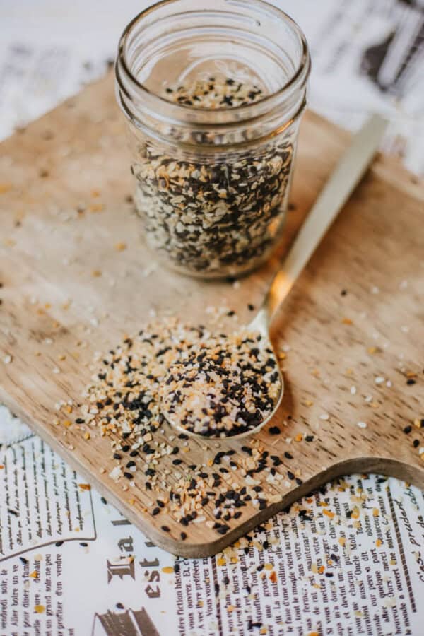 A glass jar filled with homemade everything bagel seasoning—a mix of black, white, and brown seeds—sits on a wooden board. Some seeds spill onto the board and a spoon, with more seasoning in it, rests beside the jar. The background features newspaper print.
