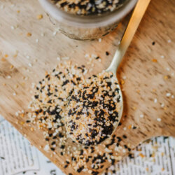 A close-up of a spoon on a wooden board, filled with homemade everything bagel seasoning—black and white sesame seeds mixed with dried onion and garlic flakes. The mixture spills onto the board, where a jar and a newspaper lie underneath.