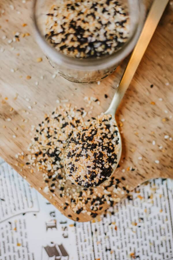 A close-up of a spoon on a wooden board, filled with homemade everything bagel seasoning—black and white sesame seeds mixed with dried onion and garlic flakes. The mixture spills onto the board, where a jar and a newspaper lie underneath.