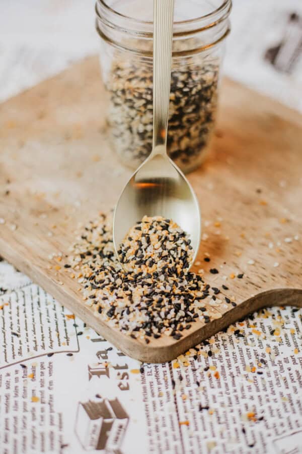 A spoon filled with black and white sesame seeds rests on a wooden board, with more seeds spilled around. A glass jar of homemade everything bagel seasoning, featuring similar seeds, is in the background on a newspaper-covered surface.