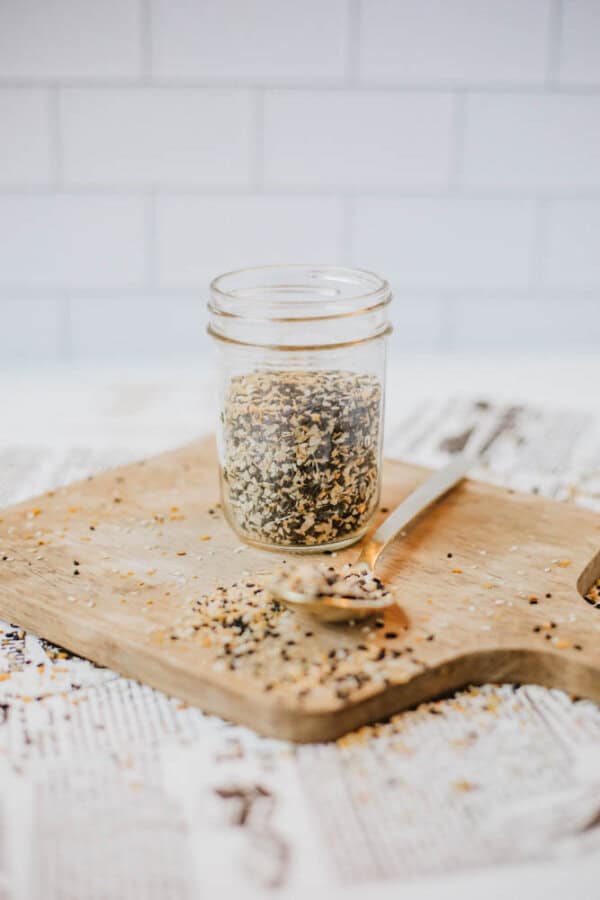 A mason jar filled with a mixed seed blend reminiscent of homemade everything bagel seasoning sits on a wooden cutting board. A wooden spoon with seeds is placed nearby, some scattered around. The background features white subway tiles and newspaper, adding a cozy kitchen vibe.