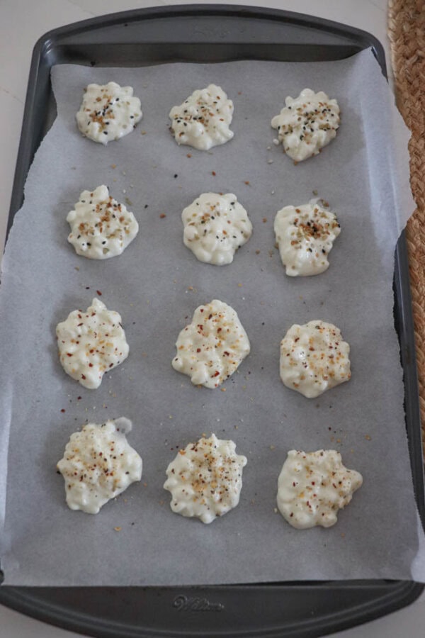 A baking tray lined with parchment paper holds twelve mounds of seasoned white batter, resembling cottage cheese. Each mound is topped with a sprinkling of mixed spices, ready to be baked into delicious chips.
