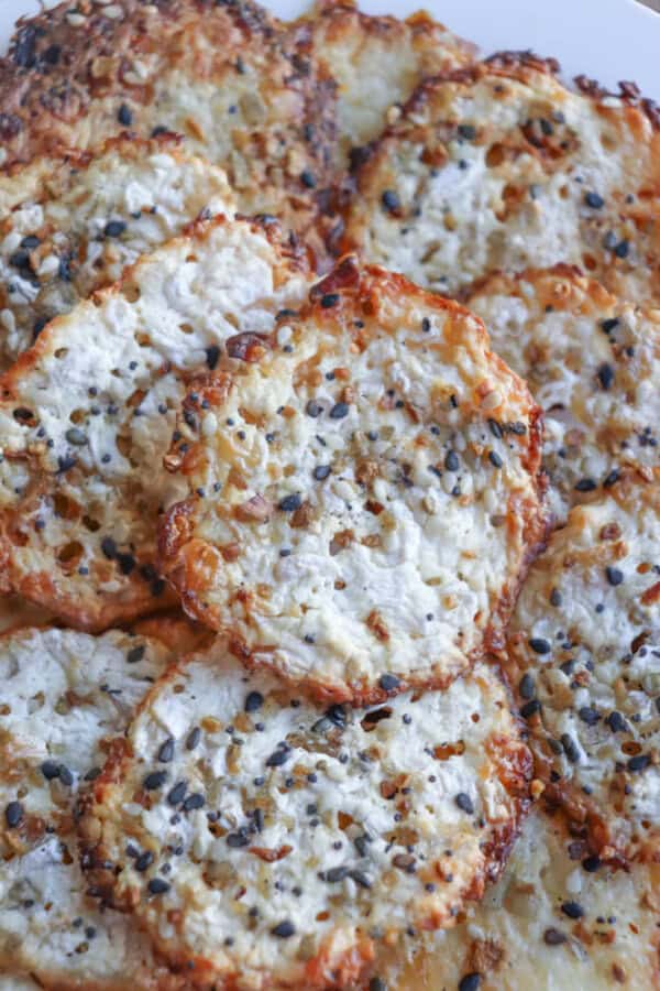 A close-up of multiple round cheesy cottage cheese chips topped with sesame seeds and seasonings, arranged on a white plate. The crisps boast a golden-brown, crispy appearance with a textured surface.