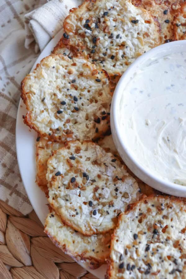 A plate of crispy round crackers topped with sesame seeds is arranged around a small bowl of creamy cottage cheese dip. The plate is set on a woven placemat next to a beige cloth.