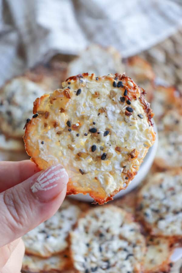 A hand holds a crispy baked snack topped with sesame seeds and spices, reminiscent of cottage cheese chips. More snacks are blurred in the background on a checkered tablecloth, while the persons fingernails display a white and pink pattern.