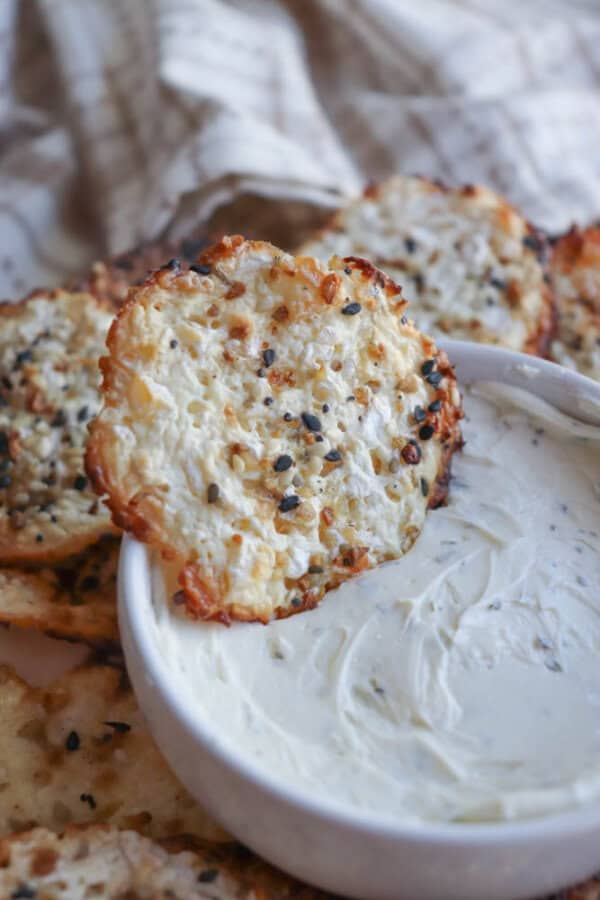 Crispy cottage cheese chips with sesame seeds and spices are arranged around a white bowl filled with creamy dip. A plaid cloth is draped in the background.