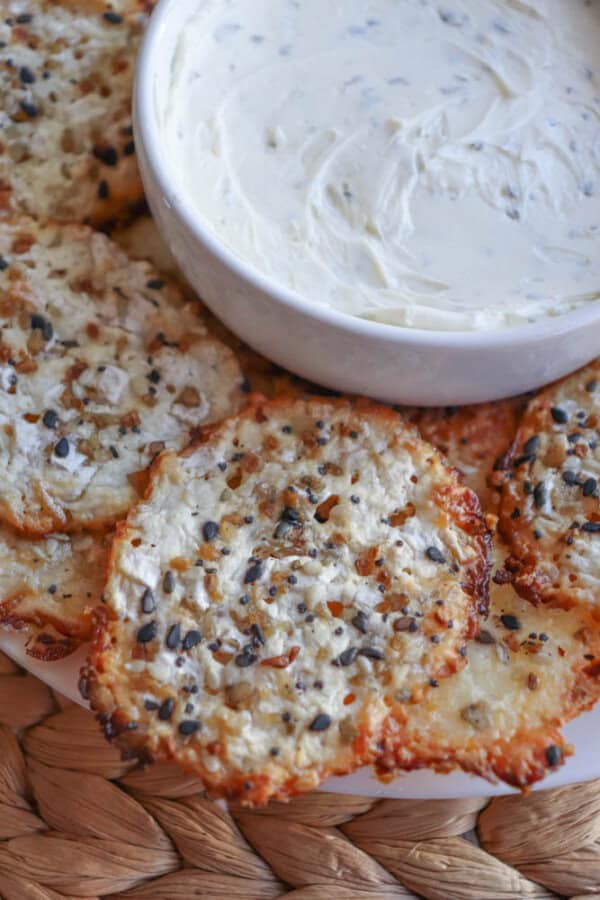 A bowl of creamy dip is surrounded by crispy cheese crackers and cottage cheese chips, all topped with seeds. The setup rests on a woven placemat, creating a rustic look.