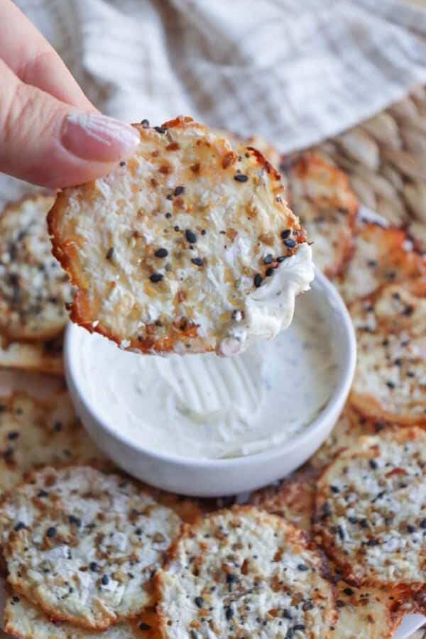 A hand holding a crispy, seasoned cracker with cream cheese dip. The cracker is topped with sesame seeds and spices. Below, more cottage cheese chips are on a woven surface around a bowl of dip.