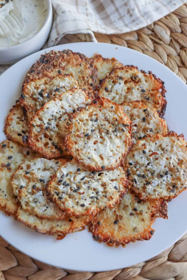 A plate of crispy cheese crisps seasoned with black and white sesame seeds, reminiscent of cottage cheese chips. The crisps are golden brown and textured, arranged in a circular pattern. A napkin and a small bowl of dip are partially visible in the background.