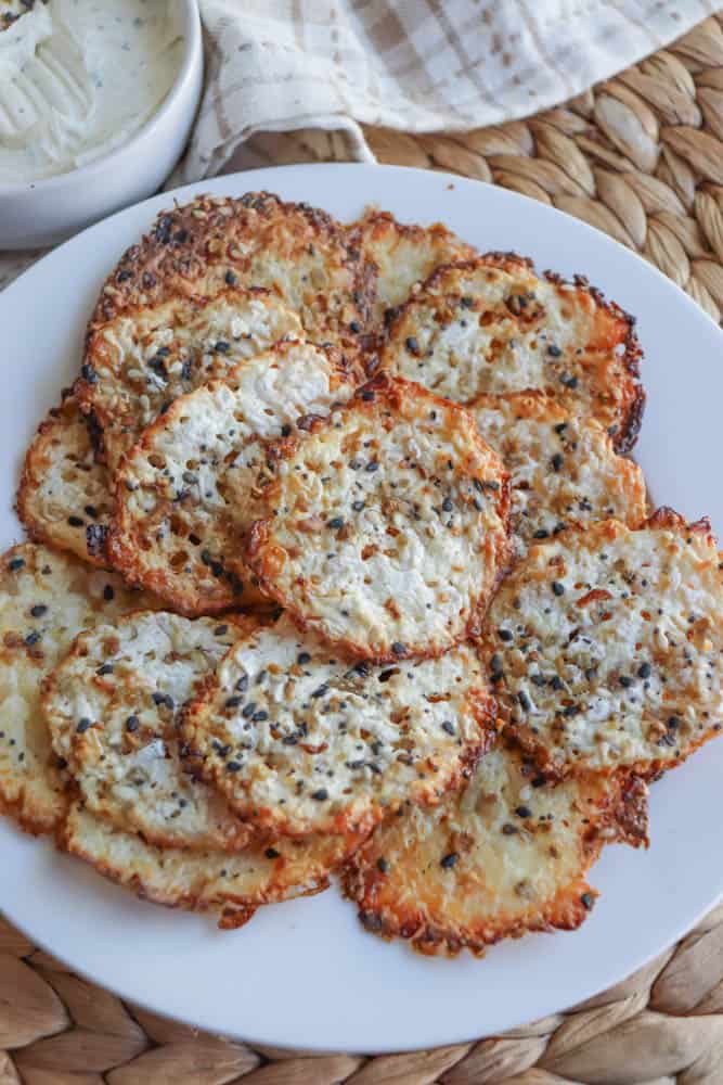 A plate of crispy cheese crisps seasoned with black and white sesame seeds, reminiscent of cottage cheese chips. The crisps are golden brown and textured, arranged in a circular pattern. A napkin and a small bowl of dip are partially visible in the background.