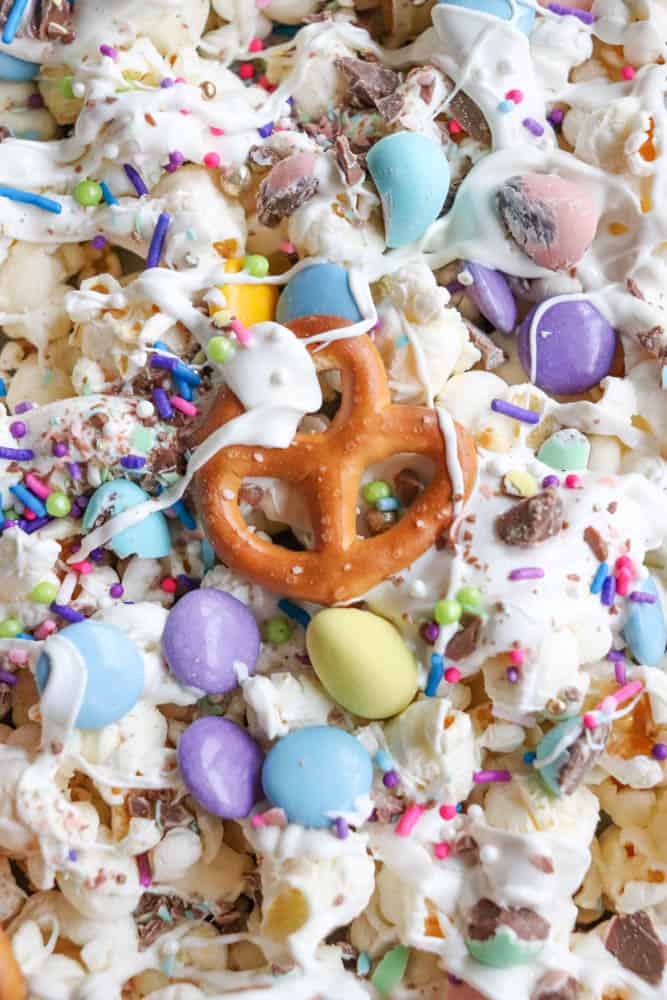 A close-up of a colorful snack mix featuring popcorn, pretzels, pastel candy-coated chocolates, and sprinkles. The mix is drizzled with white chocolate, creating a festive and vibrant treat.