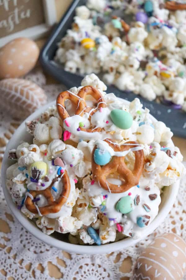 A bowl of Easter-themed snack mix featuring popcorn, mini pretzels, and candy-coated chocolates drizzled with white icing. The background has a tray with more mix and decorative eggs.