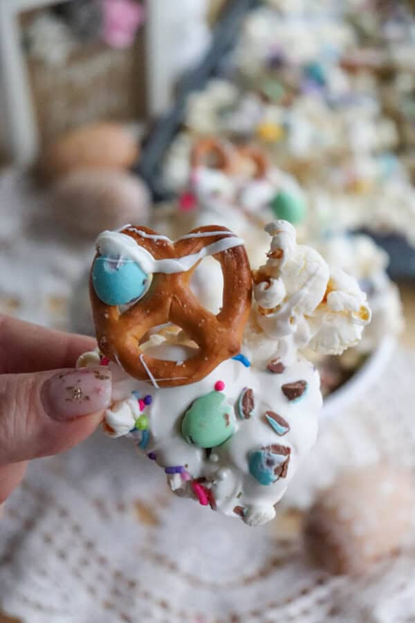 A hand holding a pretzel coated in white chocolate, topped with popcorn, colorful candy, and sprinkles. Blurred background with a tray of similar treats and a lace tablecloth.