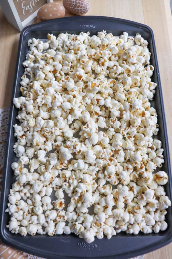 A baking sheet filled with freshly popped popcorn is placed on a wooden surface. The popcorn is evenly spread across the tray, creating a fluffy and crunchy texture. A decorative egg and a partially visible sign are in the background.
