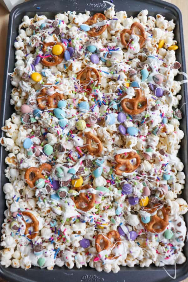 A tray of colorful Easter snack mix featuring popcorn, pretzels, and candies, drizzled with white chocolate and topped with rainbow sprinkles.