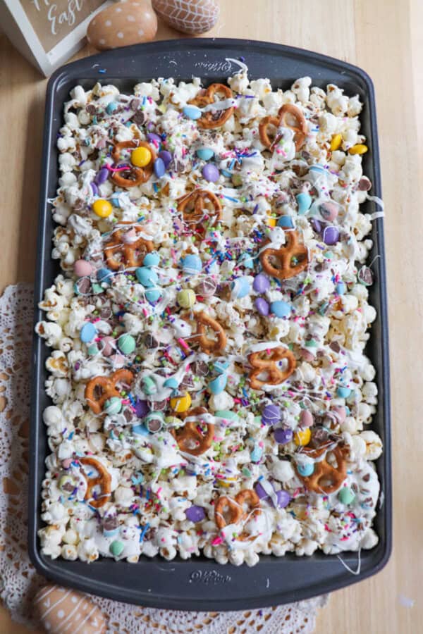 A tray filled with a colorful Easter snack mix, featuring popcorn, pretzels, pastel candies, and colorful sprinkles. It rests on a wooden table with lace doilies nearby. A painted egg is partially visible in the background.