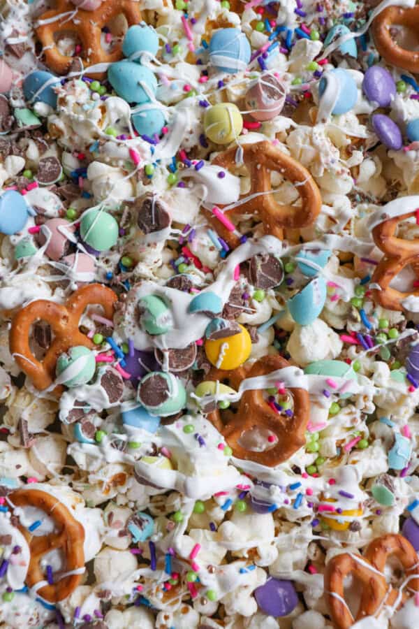 A colorful snack mix featuring pretzels, popcorn, and candy-coated chocolates drizzled with white chocolate and topped with vibrant sprinkles in shades of blue, pink, purple, and green.