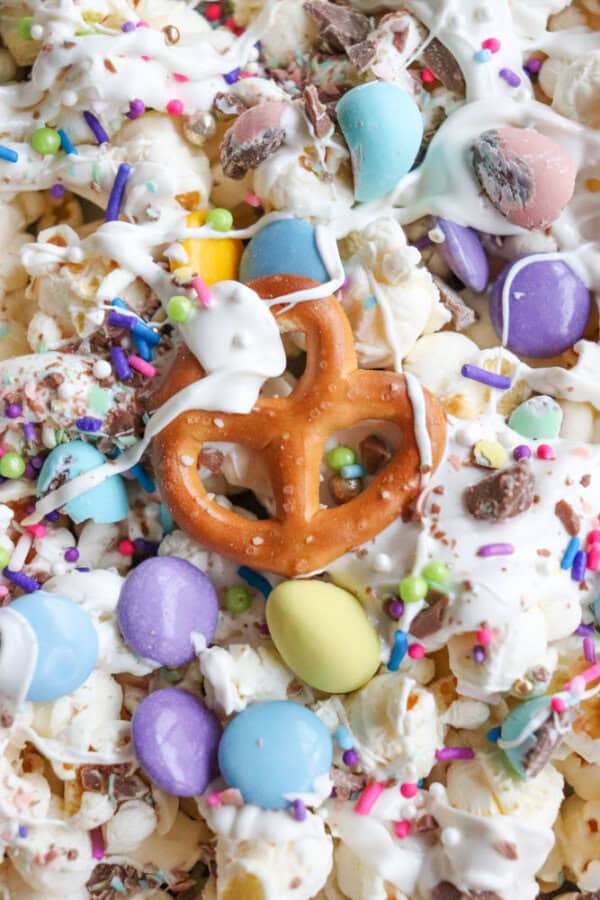 Close-up of a festive snack mix featuring popcorn, pastel candy-coated chocolates, pretzel twists, and colorful sprinkles. White chocolate drizzles bind the mixture, creating a vibrant and sweet treat.