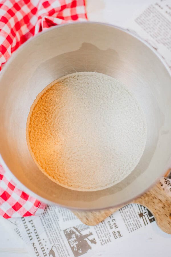 A metal bowl contains dry yeast on top of a red and white checkered cloth, ready to bring that homemade touch. Beneath the cloth are scattered newspaper clippings, hinting at a secret recipe for everything bagel seasoning. The surface below is a light, textured countertop.